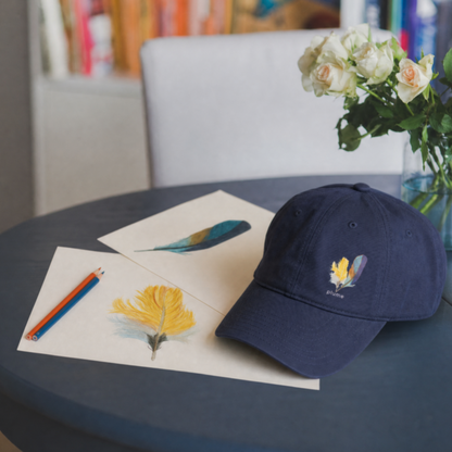 Navy blue cap with hand drawn kingfisher feathers design on a table with kingfisher feather drawing materials and flowers.