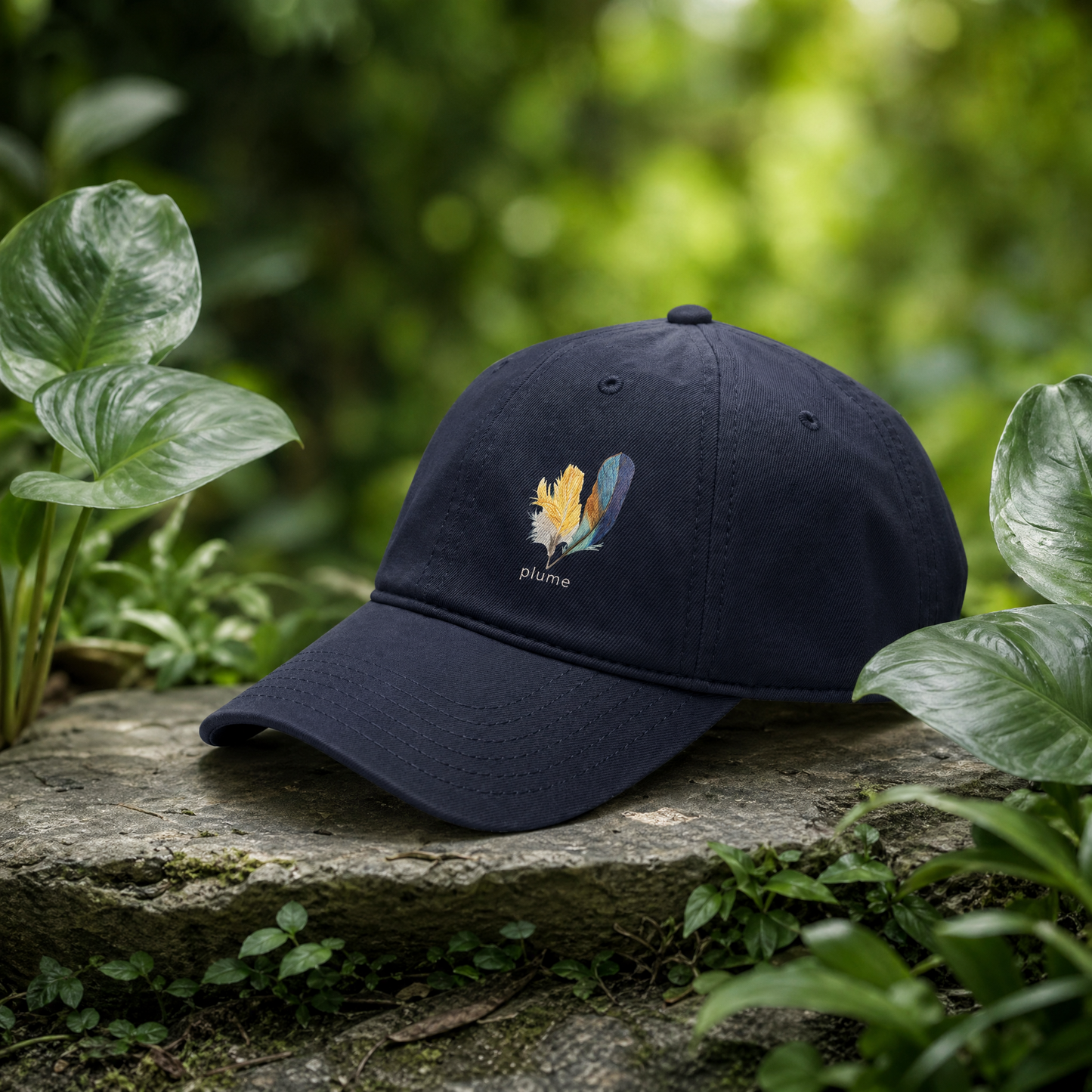 Navy blue cap with hand drawn kingfisher feather design and 'plume' branding on a stone surface with green foliage.