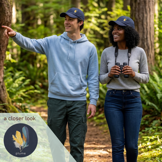 Two people in a forest, one pointing and the other holding binoculars, wearing navy dad hats with embroidered hand drawn kingfisher feathers