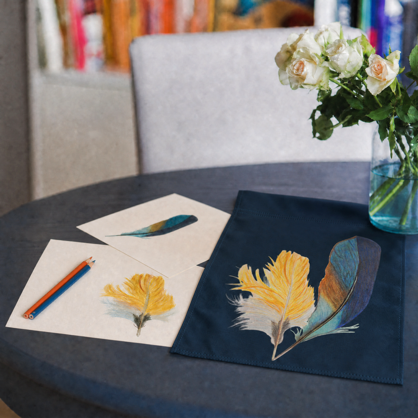Table with hand drawn kingfisher feather art on paper next to a kingfisher feather garden flag, with flowers in the background.