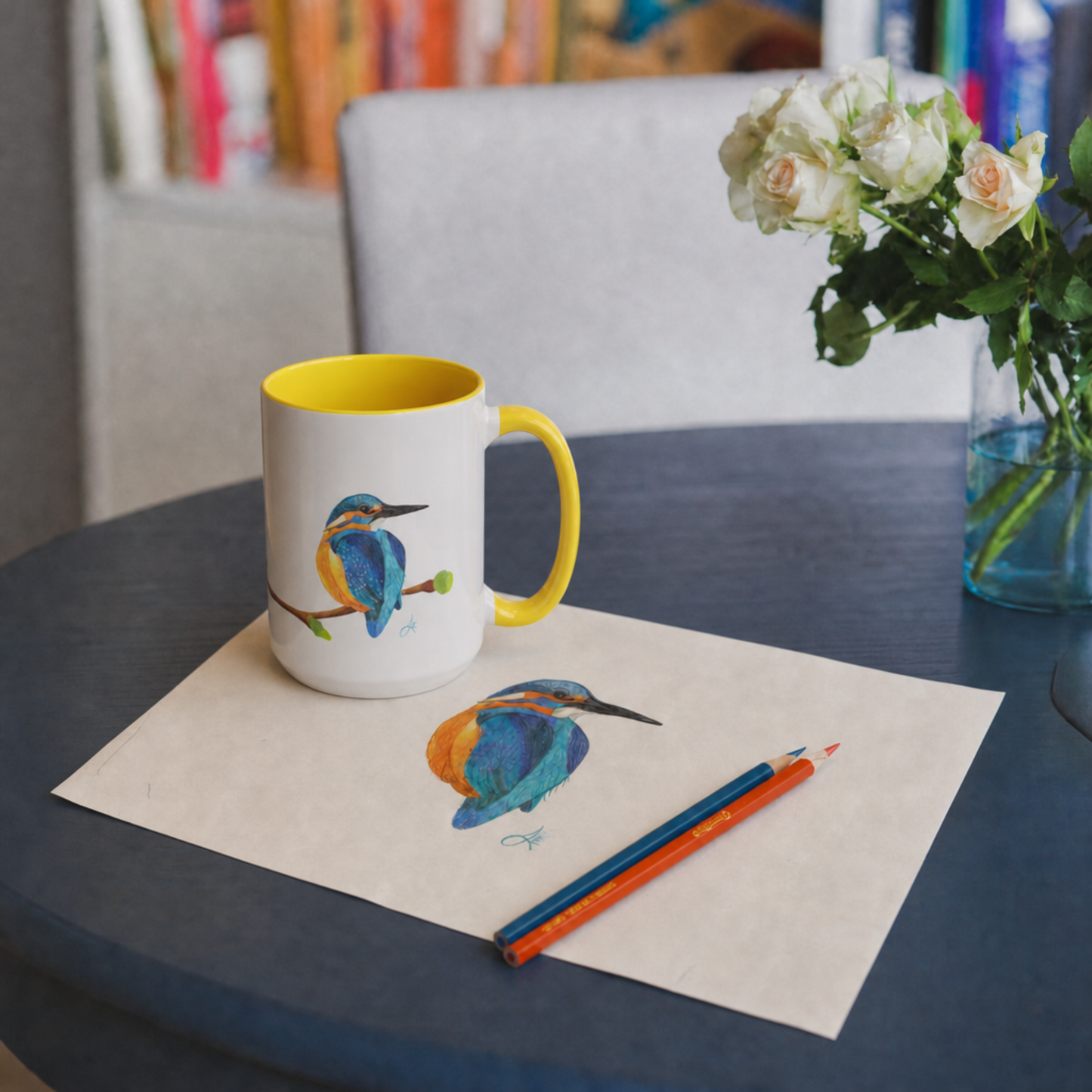 Mug with bird design on a table with paper, pencils, and flowers.