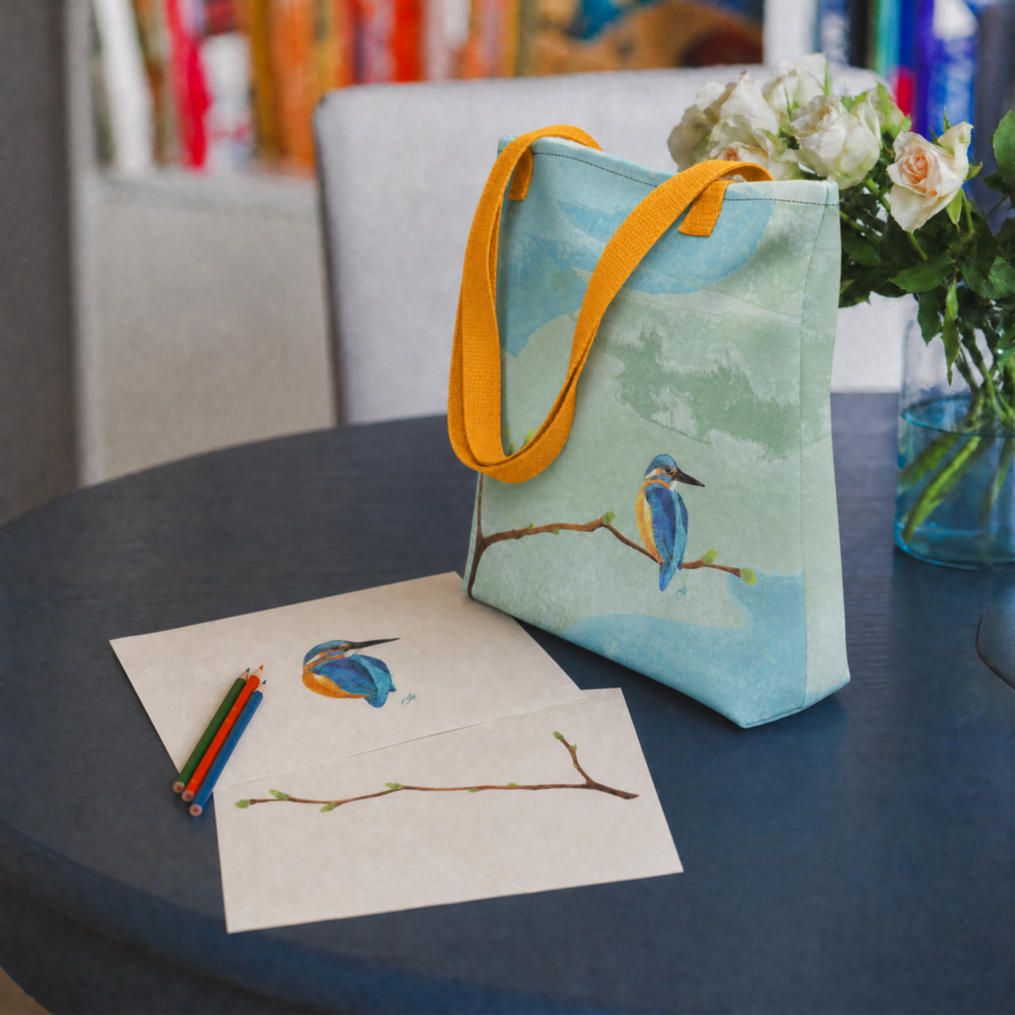 Tote bag with hand drawn kingfisher bird design on a table next to a kingfisher drawing and colored pencils