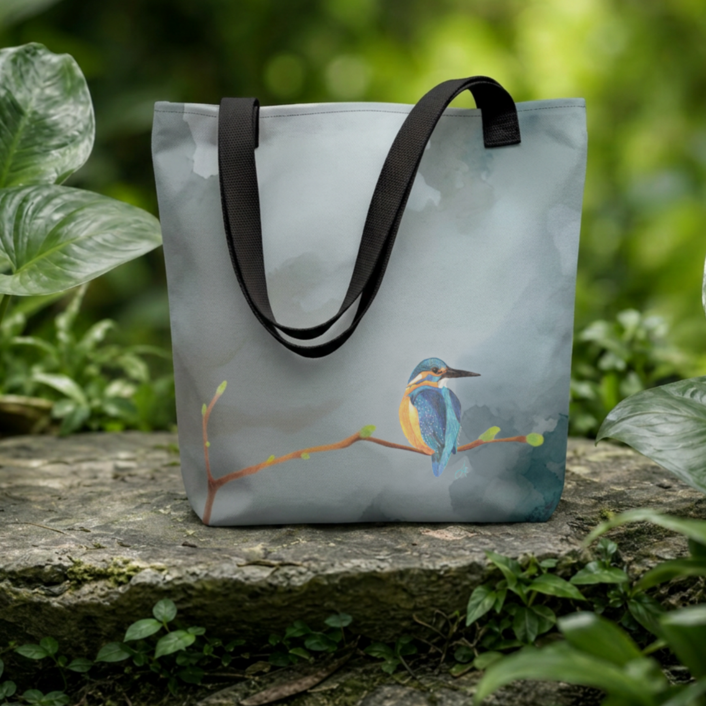 Tote bag with a hand drawn kingfisher bird design on a stone surface with greenery in the background
