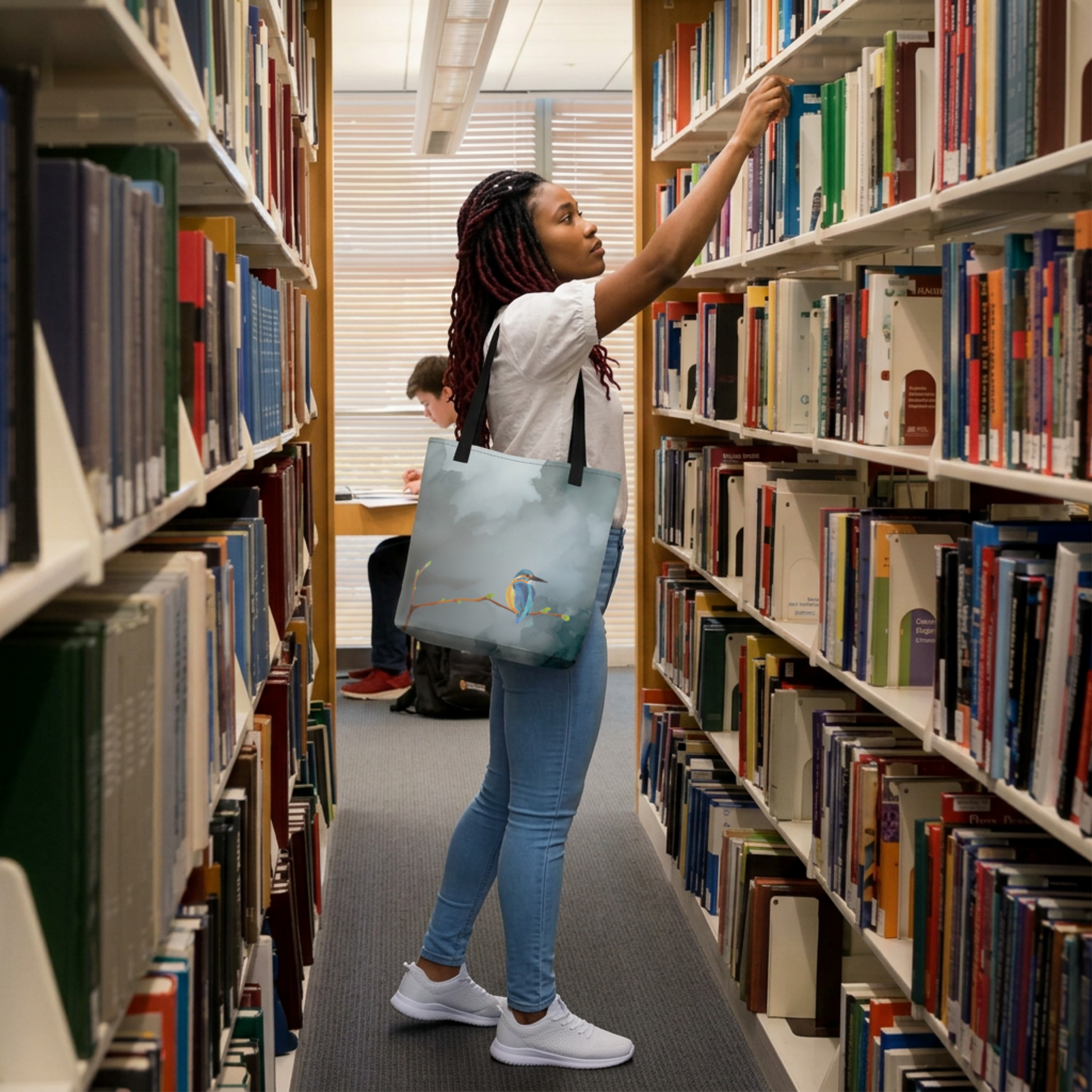 Woman browsing books in a library aisle with a hand drawn kingfisher tote bag.